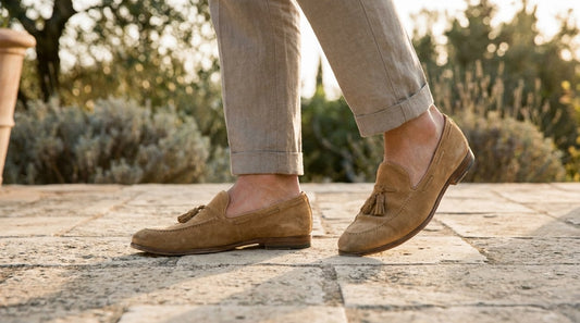 Bare ankles in tan suede loafers with cropped linen trousers on a sunlit limestone terrace